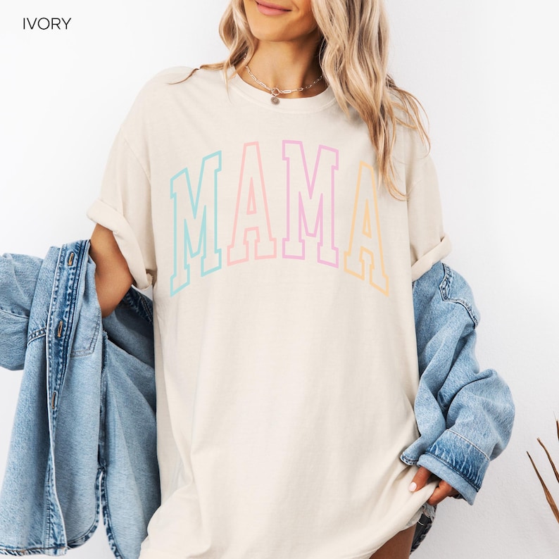May include: A light beige t-shirt with the word "MAMA" in rainbow colors. The letters are outlined in black and have a varsity style font.