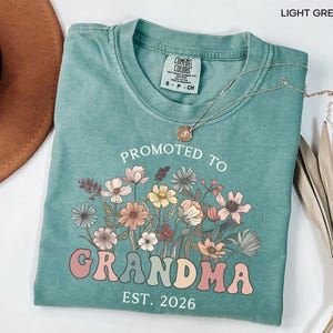 May include: Light green t-shirt with the text "PROMOTED TO GRANDMA EST. 2026" in a retro font, accompanied by a floral design in shades of pink, orange, and yellow. The shirt has a crew neck and short sleeves.