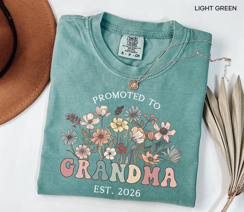 May include: Light green t-shirt with the text "PROMOTED TO GRANDMA EST. 2026" in a retro font, accompanied by a floral design in shades of pink, orange, and yellow. The shirt has a crew neck and short sleeves.