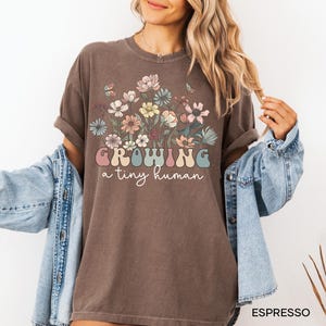 May include: Espresso-colored t-shirt with a floral design and the text "GROWING a tiny human" in a retro font. The shirt features a crew neck and short sleeves. The design includes pastel-colored flowers and butterflies.