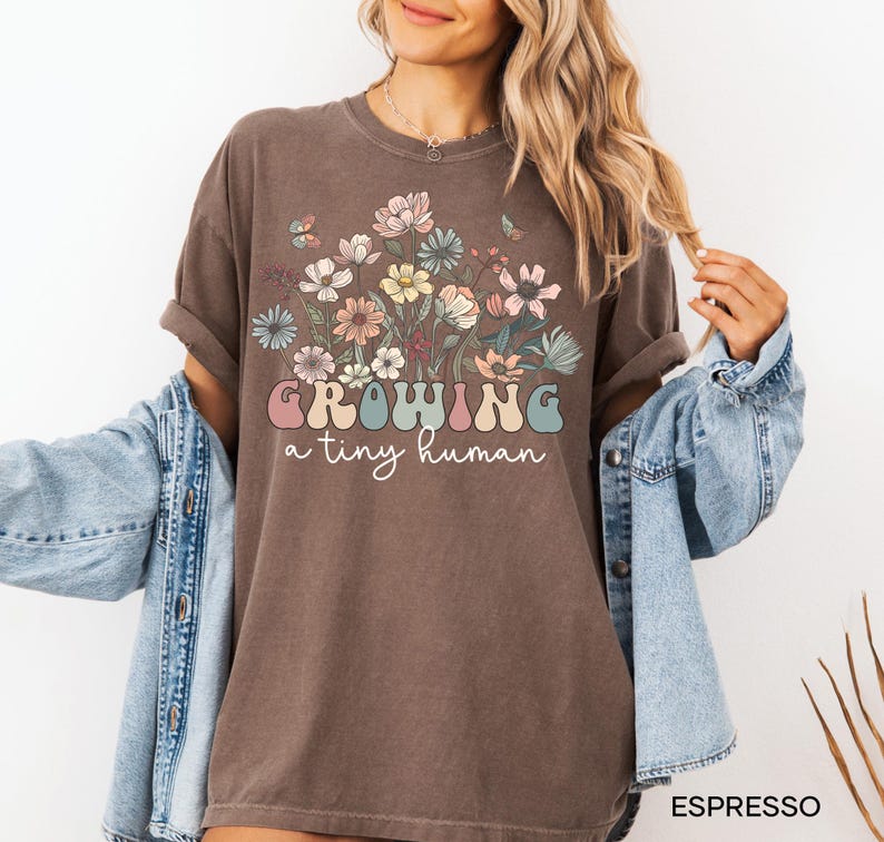 May include: Espresso-colored t-shirt with a floral design and the text "GROWING a tiny human" in a retro font. The shirt features a crew neck and short sleeves. The design includes pastel-colored flowers and butterflies.