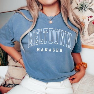 May include: A blue jean t-shirt with white text that reads "MELTDOWN MANAGER".