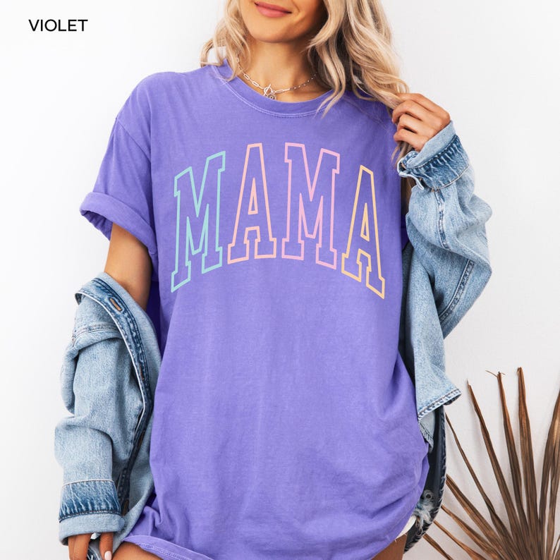 May include: A purple t-shirt with the word "MAMA" in rainbow colors. The shirt is made of a soft, comfortable material and has a relaxed fit.