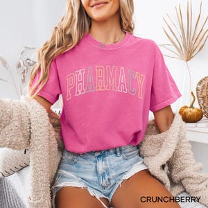 May include: A pink t-shirt with the word "PHARMACY" printed in a rainbow arch design. The shirt is worn by a person wearing denim shorts.