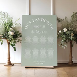 May include: A sage green wedding seating plan with white lettering. The text reads "OUR FAVOURITE PEOPLE" and "please find your seats". Names are listed under numbers one to eight. The date is 07.08.20XX. The sign is on a wooden stand.