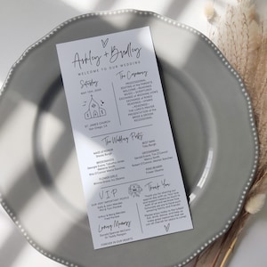 May include: A wedding program on a gray plate. The program is white with black text, including the names "Ashley + Bradley" and the wedding date. It lists the ceremony details, wedding party, and a thank you message.