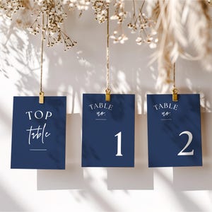 Peut inclure: Cartes de numéros de table bleu marine suspendues par des pinces dorées sur fond blanc. Les cartes affichent du texte blanc, dont "TOP Table" et les numéros 1 et 2. Des fleurs et du feuillage séchés sont visibles en haut.