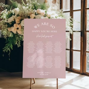 Peut inclure: Un panneau de plan de table de mariage rectangulaire rose poudré avec du texte blanc indiquant WE ARE SO HAPPY YOU'RE HERE, please find your seat, les numéros de table un à huit avec les noms des invités, TAYLOR + MARCO et 07.08.2028.