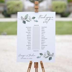Op de afbeelding: Een witte trouwzitting op een houten ezel. Het bord toont de tekst "Find your seat" in sierlijke letters, met namen en initialen van gasten. Groene en grijze bladeren versieren de boven- en onderkant. De namen Georgie & Joshua en de datum 16 oktober 2024 staan er ook op.