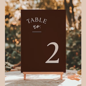 May include: A dark brown table number sign with the text "TABLE no." in a script font and the number "2" in a bold font. The sign is displayed on a small wooden easel.