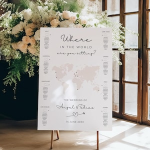 May include: A wedding seating plan on a wooden easel, with a world map and guest names. The text reads "Where in the world are you sitting?" and "The Wedding of Angel & Tobias, 14 June 20XX". The chart is decorated with a floral arrangement.