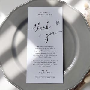Könnte beinhalten: Eine weiße Hochzeits-Dankeskarte mit eleganter Schrift, die "Danke" und ein Herz zeigt. Die Karte enthält eine Nachricht an Familie und Freunde und liegt auf einem grauen Teller.