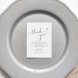 May include: A white rectangular card with the words "thank you" in elegant script, accompanied by a heart symbol. The card includes a message of gratitude and is placed on a gray plate, suitable for wedding or event favors.
