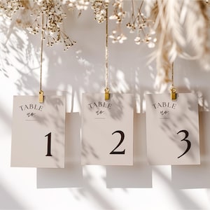 May include: Three beige table number cards suspended by gold clips and string. Each card displays "TABLE no." at the top, with the numbers 1, 2, and 3 in dark brown. Dried flowers are visible in the background.