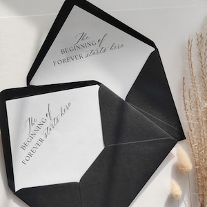 May include: Two black envelopes with white inserts. The inserts have the text "The Beginning of Forever starts here" in elegant script. The envelopes are open, revealing the text. The background is white with dried plants.
