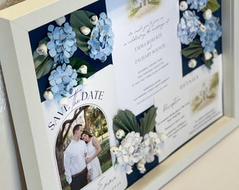 May include: A framed wedding invitation with a white border. The invitation features a photo of a couple, floral accents, and the words "Save the Date." Blue paper flowers and greenery are arranged around the invitation.