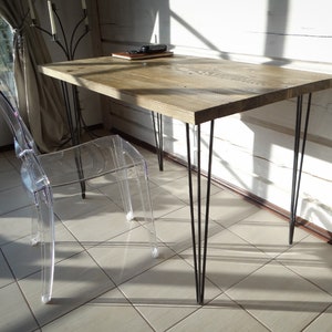 May include: A wooden desk with a natural finish and black hairpin legs. The desk is in a room with white walls and a clear plastic chair.