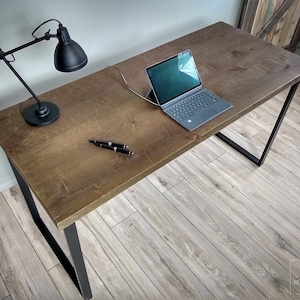 Op de afbeelding: Een rustiek houten bureau met zwarte metalen poten. Het bureau heeft een natuurlijke houten afwerking en is voorzien van een laptop, een lamp en twee pennen.