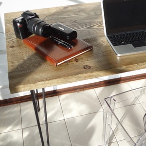 May include: A rustic wooden desk with black hairpin legs and a clear plastic chair. A laptop, camera, and notebook are on the desk.