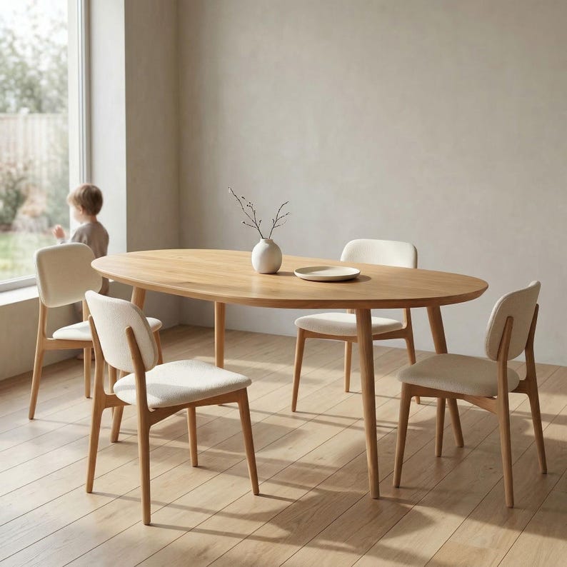 May include: An oval wooden dining table with four matching chairs. The table is light brown and has a smooth surface. The chairs have light-colored upholstered seats and backs. A small white vase and plate are on the table.