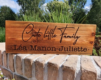 以下が含まれることがあります： 「Cute little family」というテキストと、「Léa、Manon、Juliette」という名前が黒で刻まれた木製の看板です。