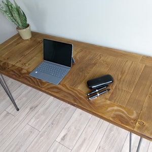 Könnte beinhalten: Ein rustikaler Holzschreibtisch mit einer natürlichen Holzoptik. Der Schreibtisch hat einen Laptop, ein schwarzes Stiftetui und zwei Stifte auf der Oberfläche. Der Schreibtisch wird von Haarnadelbeinen getragen.