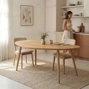 May include: A light wood oval dining table with three matching chairs. The chairs have beige upholstered seats and light wood legs. A beige rug is under the table and chairs. A vase of flowers sits on the table. A woman is in the background.