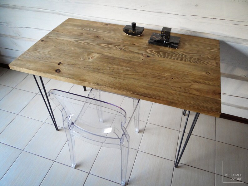 May include: A rustic wooden desk with black hairpin legs and a clear plastic chair. The desk has a natural wood finish and is made from reclaimed wood. The chair is a modern design and is made from clear acrylic.