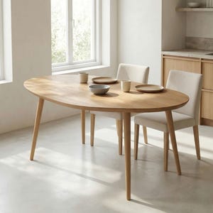 May include: An oval wooden dining table with four angled legs, set with plates, bowls, and cups. Two upholstered chairs with light-colored fabric are positioned around the table. The room has a neutral color palette with natural light.