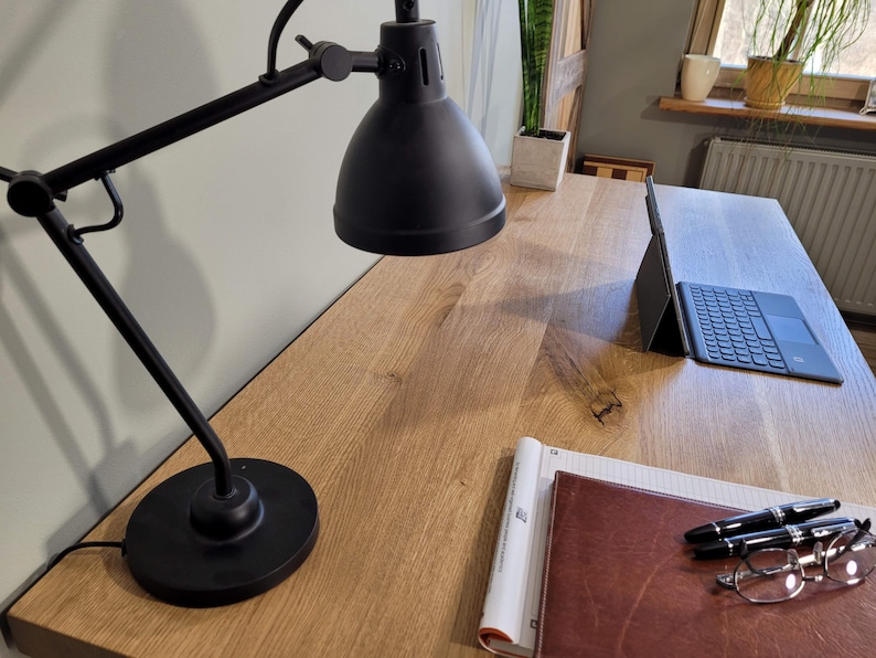 May include: A wooden desk with a black desk lamp, a laptop, a notebook, a leather-bound journal, two pens, and a pair of glasses.
