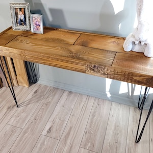 May include: A rustic wooden console table with a natural wood finish. The table has a rectangular top and is supported by four black hairpin legs. The table is decorated with two framed pictures and a white ceramic rabbit figurine.