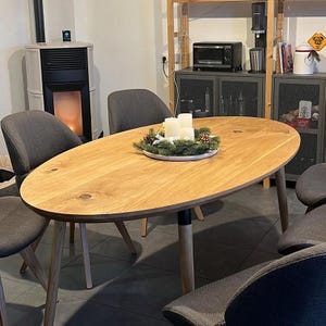 Op de afbeelding: Een ovale eettafel van licht hout met een zwarte basis en vier grijze stoelen. De tafel is versierd met een groene krans en witte kaarsen.