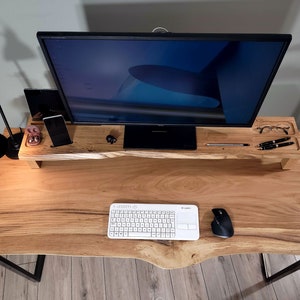Könnte beinhalten: Ein Holztisch mit einer Live-Edge-Oberfläche und schwarzen Metallbeinen. Ein Monitor steht auf einem Holzständer mit einer Tastatur und einer Maus auf dem Tisch. Auf dem Ständer befinden sich verschiedene Accessoires, darunter ein Telefon, Ohrhörer und ein Stift.