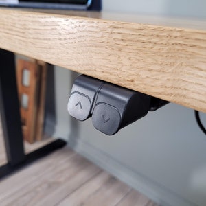 May include: Close-up of a black electric standing desk control panel with two buttons, one with an up arrow and one with a down arrow. The control panel is mounted under a wooden desk.