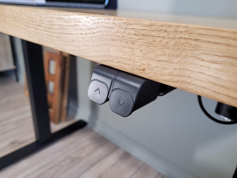 May include: Close-up of a black electric standing desk control panel with two buttons, one with an up arrow and one with a down arrow. The control panel is mounted under a wooden desk.