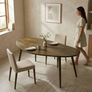 May include: Oval wooden dining table with four legs, set with plates, cups, and a vase of flowers. Two upholstered chairs are positioned around the table. A woman in a dress walks past the table in a well-lit room.