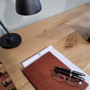 May include: A wooden desk with a black lamp, a brown leather notebook, two pens, a pair of glasses, and a laptop computer.