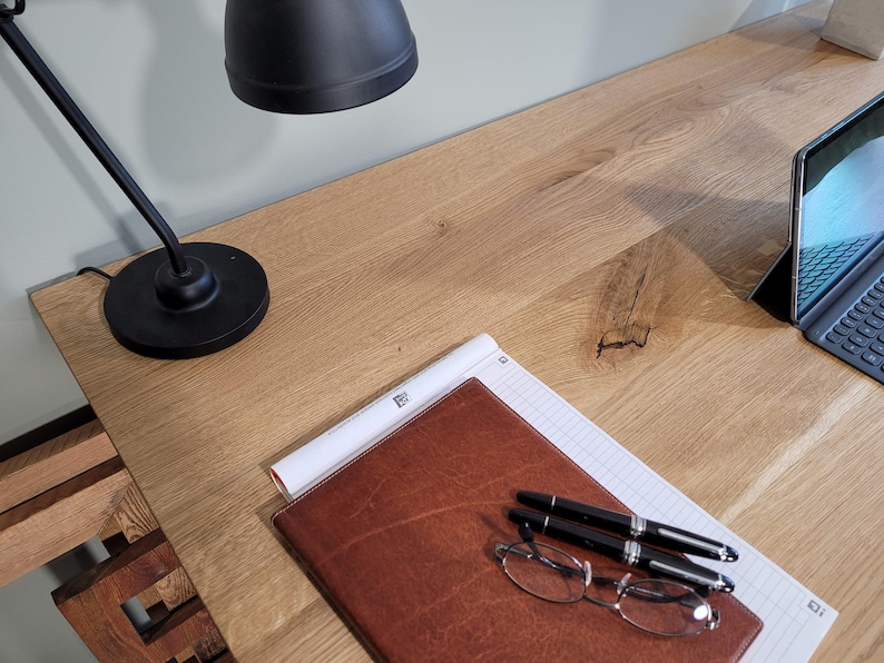 May include: A wooden desk with a black lamp, a brown leather notebook, two pens, a pair of glasses, and a laptop computer.