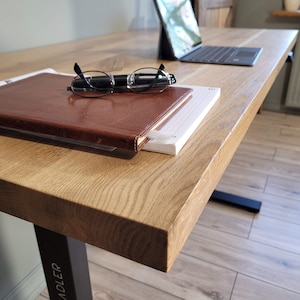 May include: A wooden desk with a brown leather-bound journal, a pair of glasses, and a pen on top. The desk has a black metal leg with the word "ADLER" printed on it.