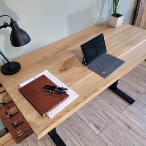 May include: A wooden desk with a black metal frame and a laptop computer, a notebook, pens, and glasses on the surface. A black desk lamp is on the left side of the desk.