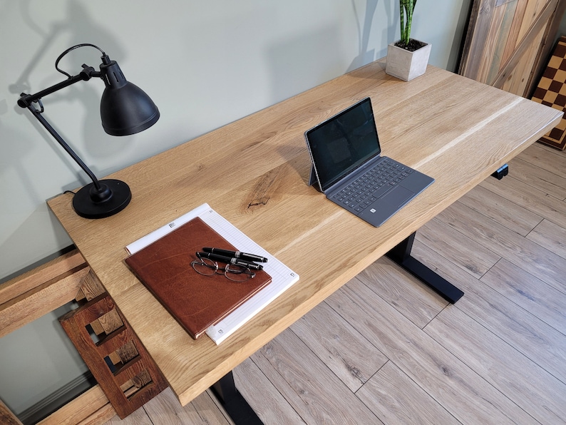 May include: A wooden desk with a black metal frame and a laptop computer, a notebook, pens, and glasses on the surface. A black desk lamp is on the left side of the desk.