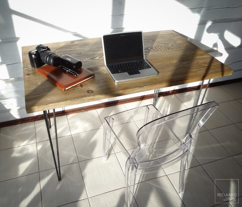 May include: A rustic wooden desk with black hairpin legs and a clear plastic chair. A laptop, camera, and notebook are on the desk.