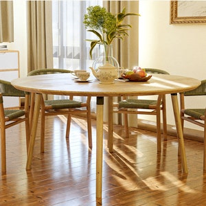 May include: A round wooden dining table with four chairs. The table has a light wood finish and the chairs have woven green seats and backs. There are green plants in a vase in the center of the table, along with a teapot and cups.