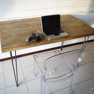 May include: A rustic wooden desk with black hairpin legs and a clear plastic chair. A laptop computer and a black case are on the desk.