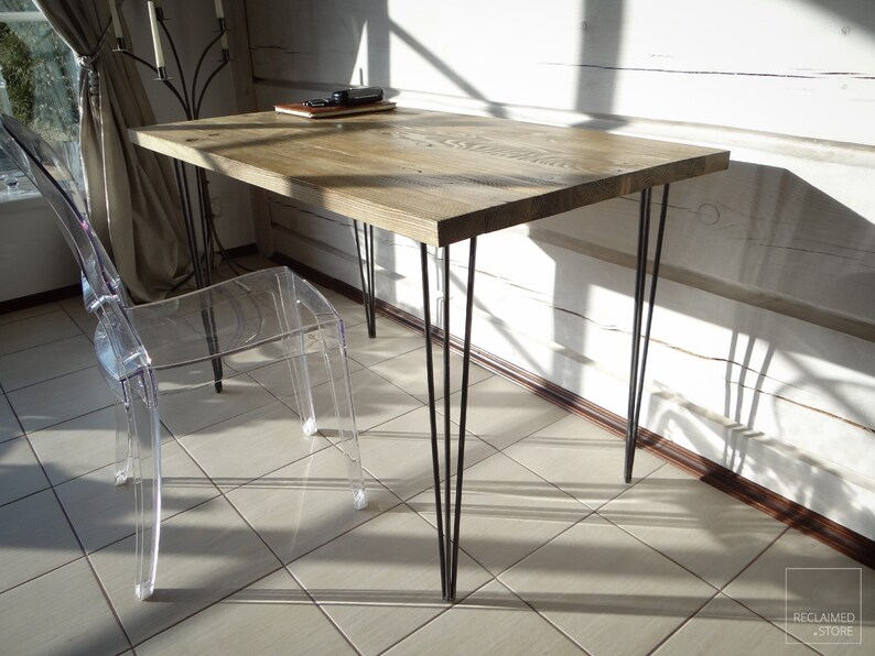 May include: A wooden desk with a natural finish and black hairpin legs. The desk is in a room with white walls and a clear plastic chair.
