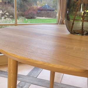 May include: A close-up of a round, wooden table top with a smooth, light brown finish. The table has a slightly rounded edge and is made of solid wood.