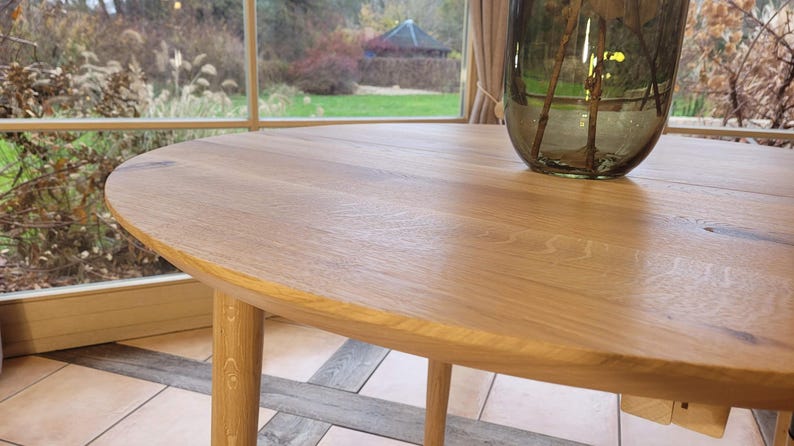 May include: A close-up of a round, wooden table top with a smooth, light brown finish. The table has a slightly rounded edge and is made of solid wood.