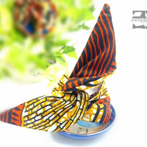 Puede incluir: Una diadema de tela colorida con un diseño anudado. La diadema está hecha de una tela estampada con colores naranja, amarillo, negro y blanco. La diadema está sobre un plato azul y blanco.