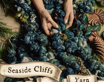 Pode incluir: Um close-up de uma mão segurando um novelo de lã azul e verde. A lã é chamada de "Seaside Cliffs Yarn".