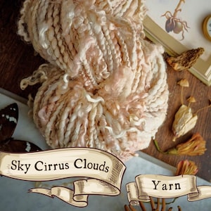 Puede incluir: Un gran ovillo de hilo Sky Cirrus Clouds, con una mezcla de fibras crema y rosa pálido. El hilo tiene un aspecto texturizado y voluminoso, ideal para proyectos de tejido o ganchillo. La palabra "Yarn" también es visible.